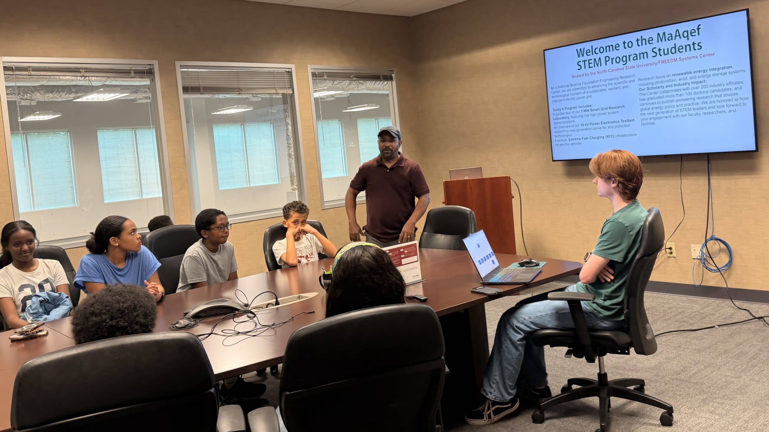 Speaker standing in front of screen presenting to students seated at a large conference table.